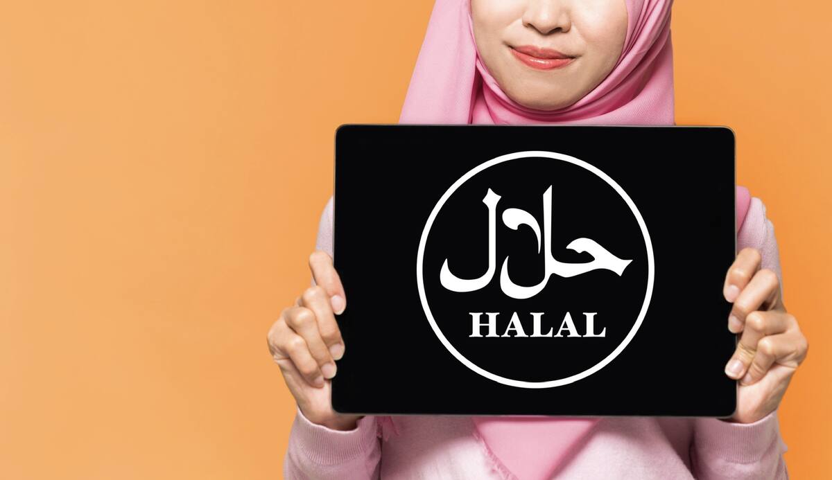 Woman in a pink hijab holding up a tablet displaying a white halal symbol and the word halal in Arabic and English
