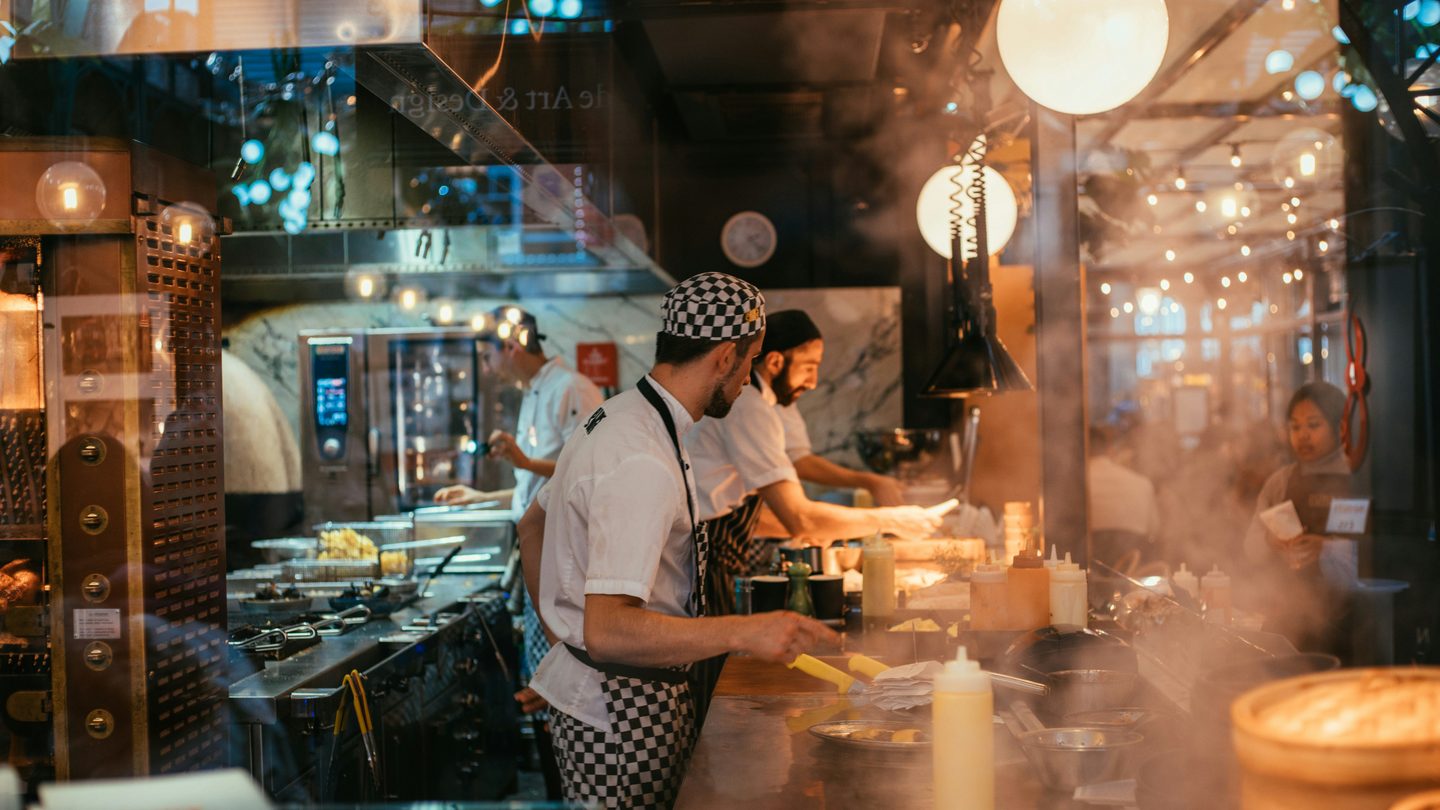 Cooks working a busy restaurant line behind glass, with fryer baskets, squeeze bottles and steam rising over the pass