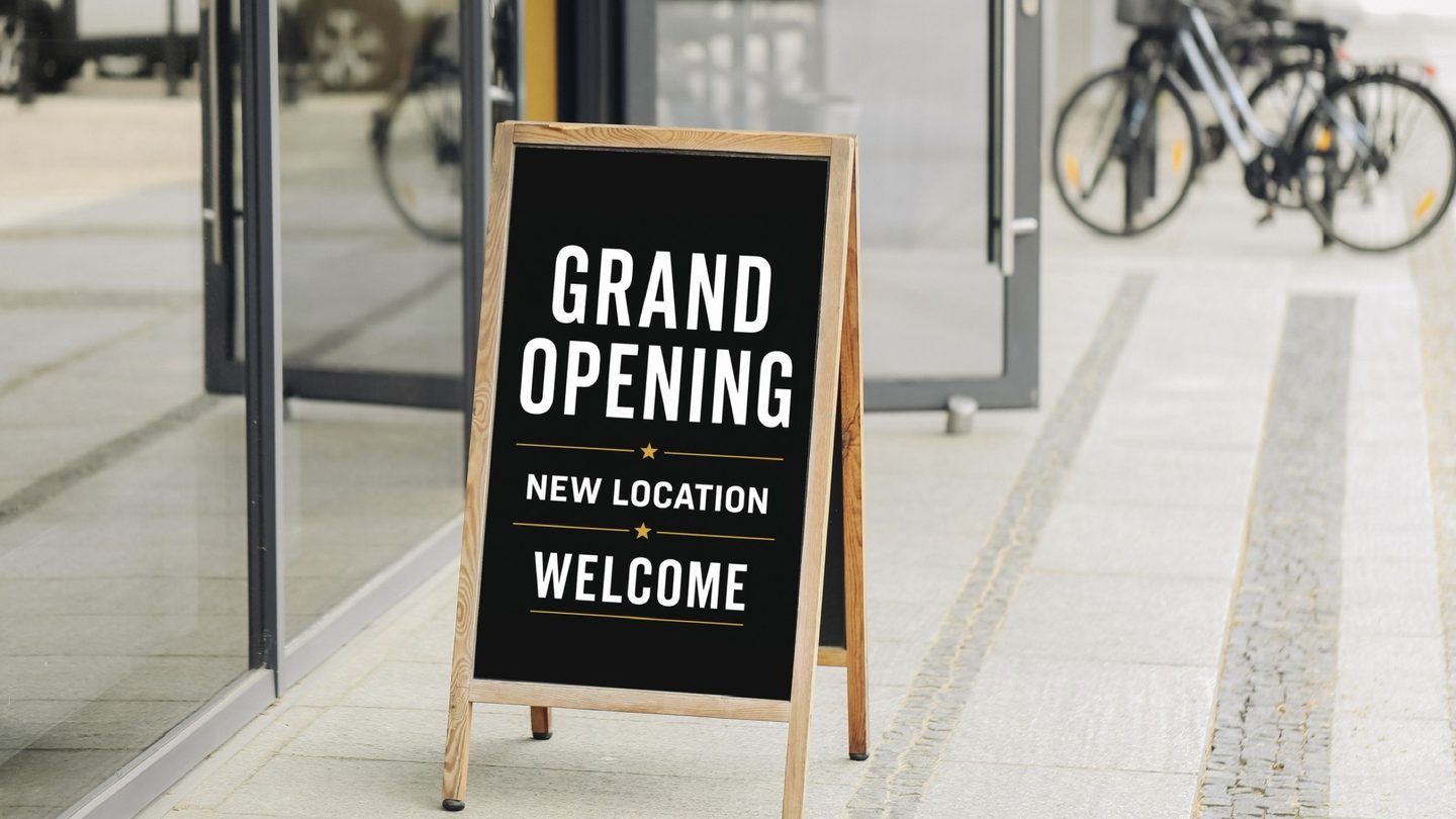 A folding "Grand Opening, New Location, Welcome" sign on the pavement outside a newly fitted storefront.