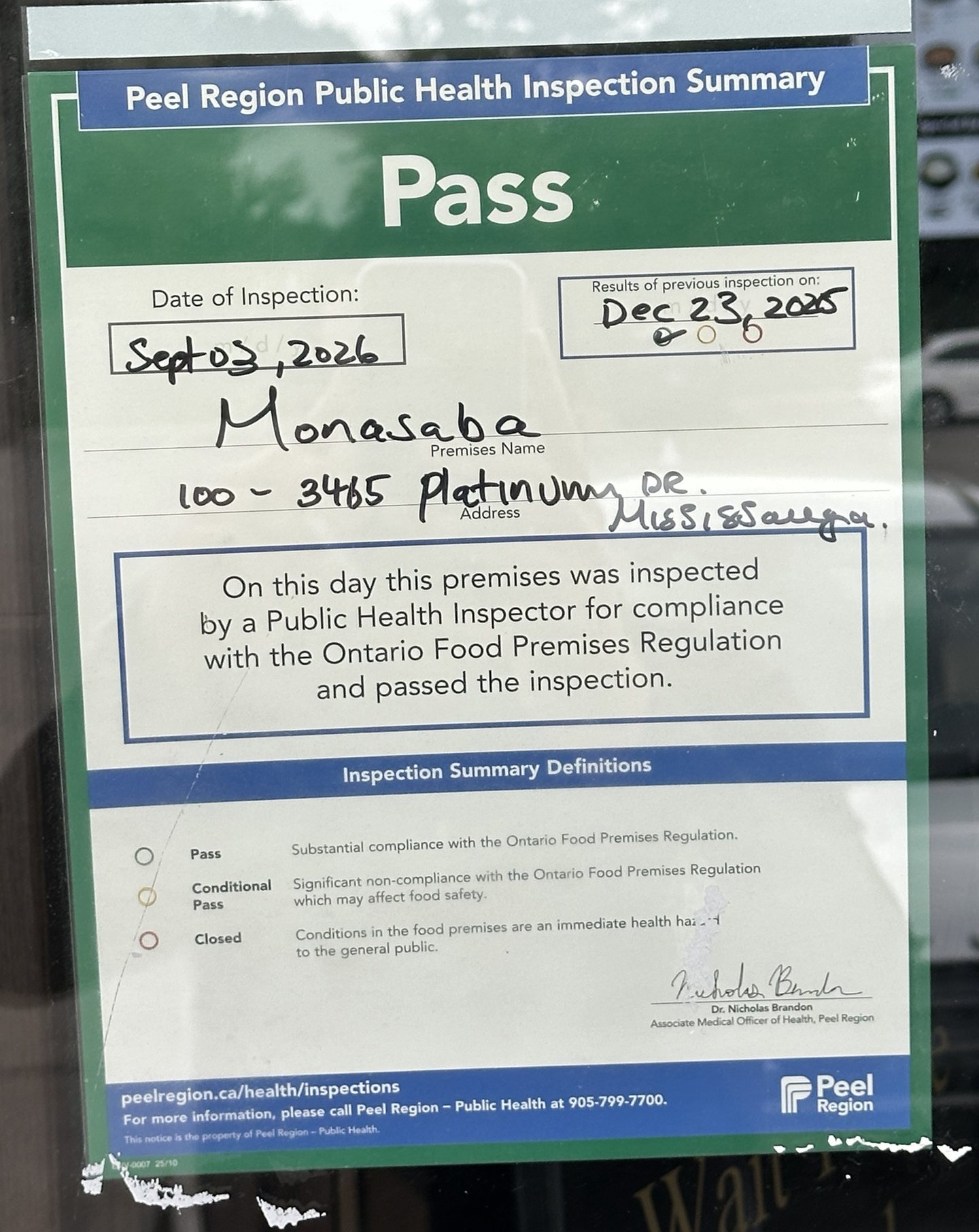 Peel Region Public Health Inspection Summary notice reading Pass, dated Sept 03, 2026, premises name Monasaba, address 100 - 3465 Platinum Dr, Mississauga, with the previous inspection recorded as Dec 23, 2025, photographed through the restaurant door glass